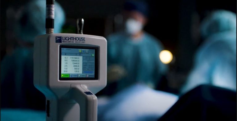 A photograph of the LWS Handheld particle counter in a dimly lit room with surgerons and nurses blurred in the background.