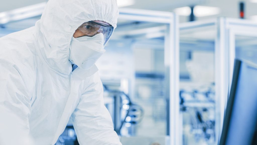 A photograph of a gowned man in a cleanroom environment looking over readings on a computer monitor.